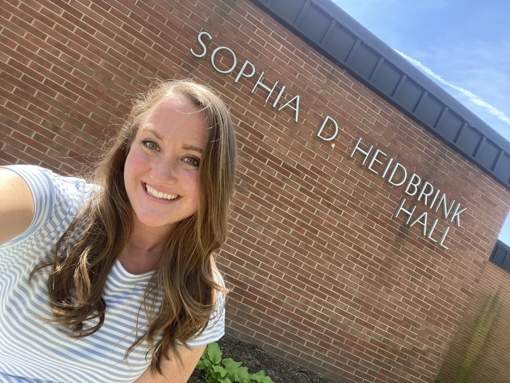 Woman smiling in front of a brick building labeled "Sophia D. Heidbrink Hall" on a sunny day, with Beacons of sunlight highlighting the scene—possibly Nicole Moy enjoying her backyard moment.