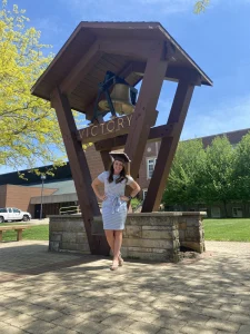 Nicole Moy, wearing a graduation cap, poses by the large "VICTORY" bell structure on a sunny day—a true beacon in your backyard.