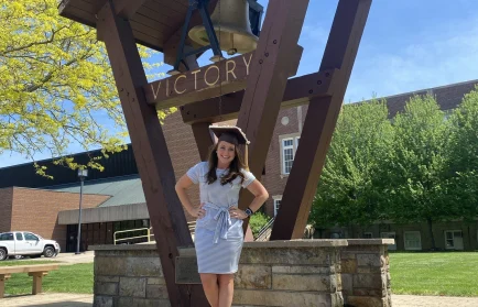 Nicole Moy, wearing a graduation cap, poses by the large "VICTORY" bell structure on a sunny day—a true beacon in your backyard.