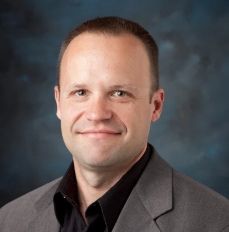 Man in a gray suit jacket and black shirt smiling in front of a dark blue background, showing support for Ukrainian Refugees.