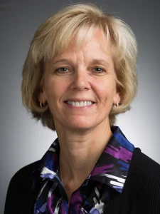 Smiling woman with short blonde hair, wearing a patterned shirt and black sweater, stands against a gray background—ready to tackle any microscopic problem or take on her next big grant.