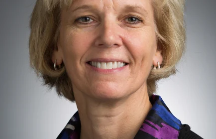 Smiling woman with short blonde hair, wearing a patterned shirt and black sweater, stands against a gray background—ready to tackle any microscopic problem or take on her next big grant.