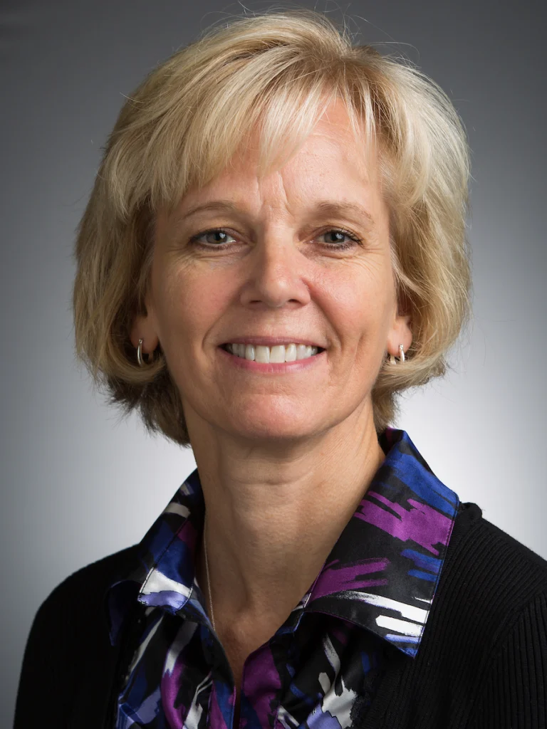 Smiling woman with short blonde hair, wearing a patterned shirt and black sweater, stands against a gray background—ready to tackle any microscopic problem or take on her next big grant.