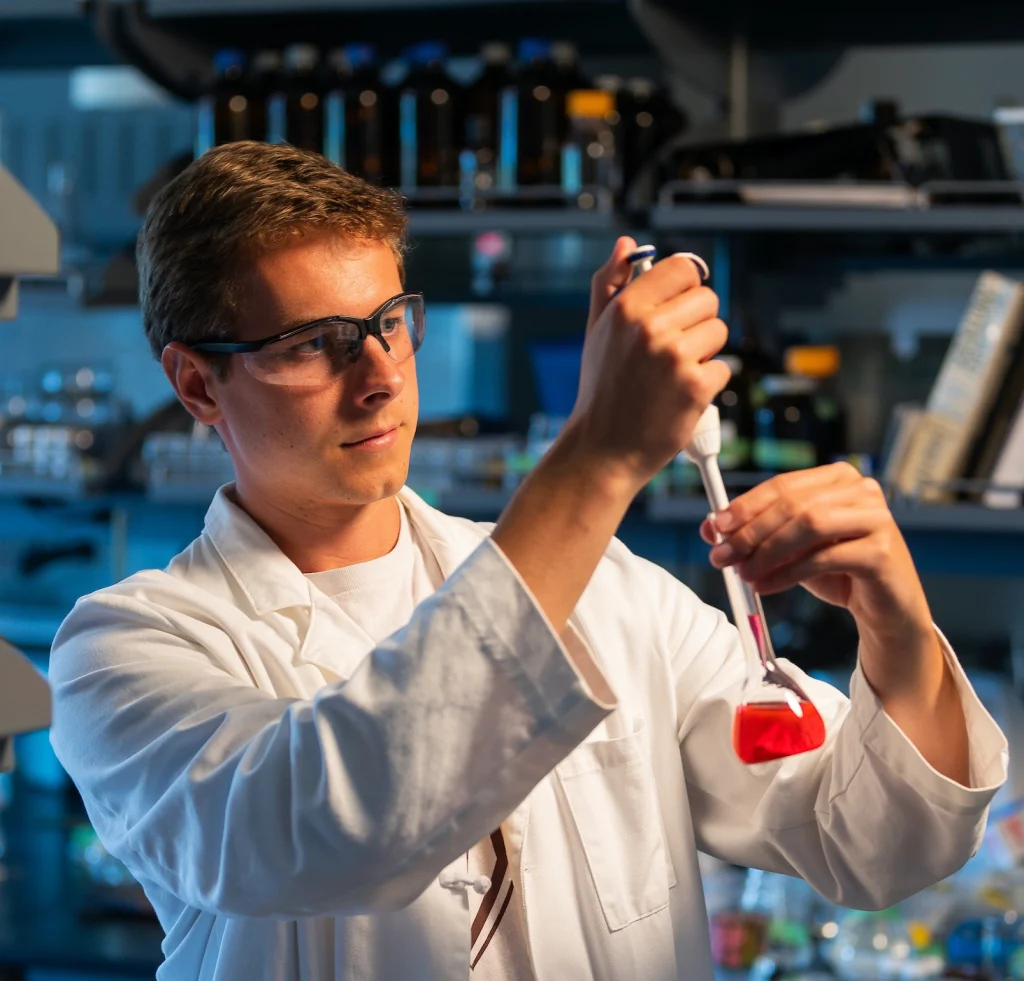 A scientist in a lab coat uses a pipette to transfer red liquid, working to solve a microscopic problem in a laboratory setting.