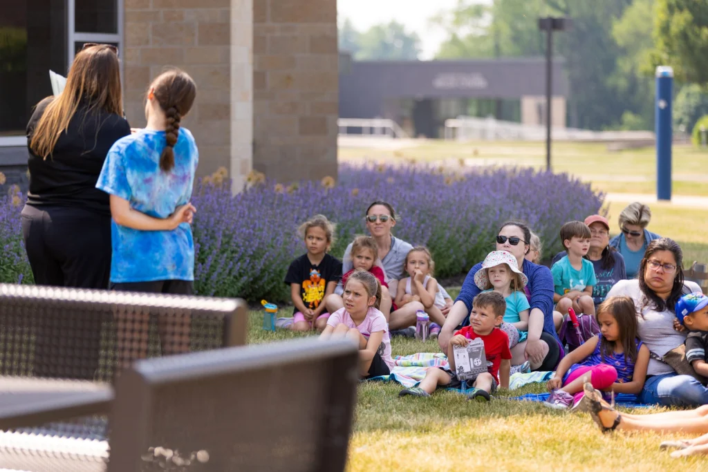 Children sit on the grass outside, enjoying Nature Play Day as they listen to two women standing and reading to them on a sunny afternoon.