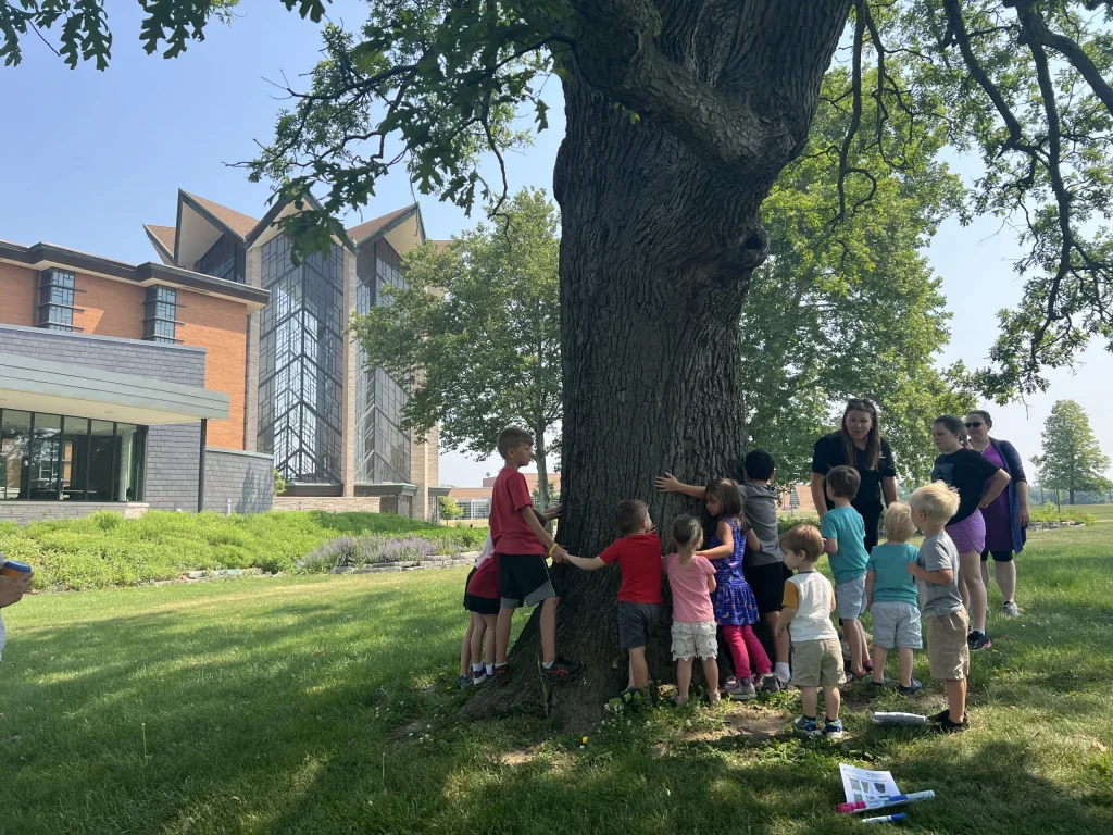 A group of children and adults stand around a large tree on a grassy lawn near a modern building, enjoying an Inclusive Nature Play day together.