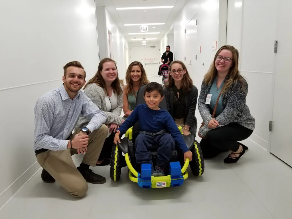 Five adults smile and crouch around a young boy in a wheelchair in a bright hallway, celebrating the launching of Valpo’s GoBabyGo initiative.
