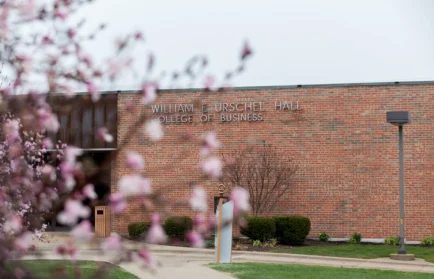 Brick building labeled "William F. Urschel Hall College of Business" at Valparaiso University, with a pink-blossomed tree in the foreground—a welcoming setting for MBA students at a Best University.