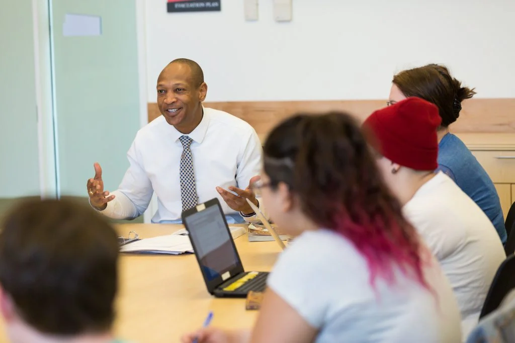 A Valpo professor in a white shirt speaks to students seated around a table with laptops and notebooks, fostering education and discussion.