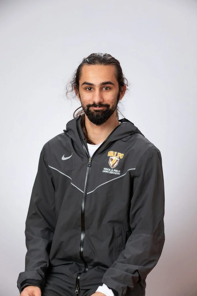 A man with long dark hair and a beard, wearing a dark athletic jacket, smiles in front of a gray background—an inspiring member of the Fulbright Scholars community at Valparaiso University.