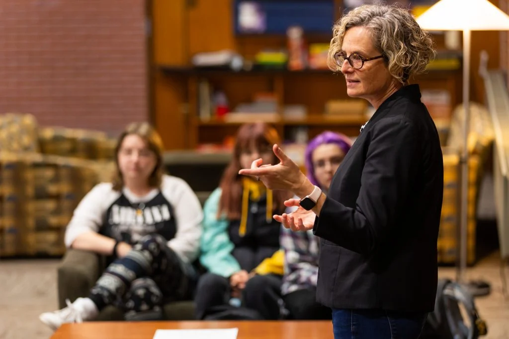 A woman with a passion for education speaks to a small seated group in a cozy room with bookshelves and warm lighting.