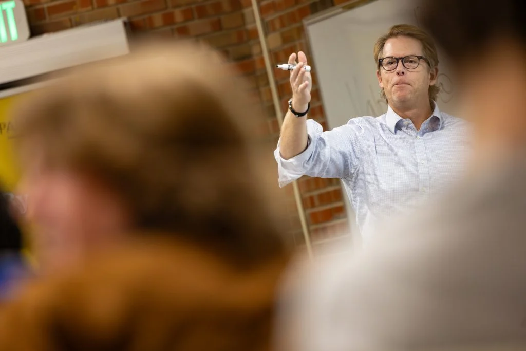 A person stands in front of a whiteboard, holding a marker and gesturing while speaking to a group about MBA programs at Valparaiso University.