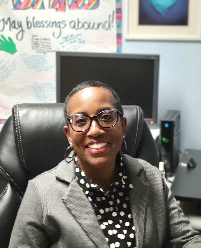 Smiling woman with glasses sits at a desk in an office, with a colorful poster behind her, welcoming new faculty to Valparaiso University's Master of Social Work program.
