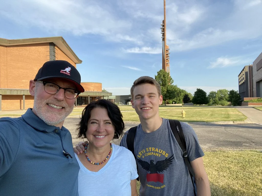 Three people, including Charles Smith, smiling outside on a sunny day, with brick buildings and a tall tower in the background, celebrating the Spirit of Innovation.