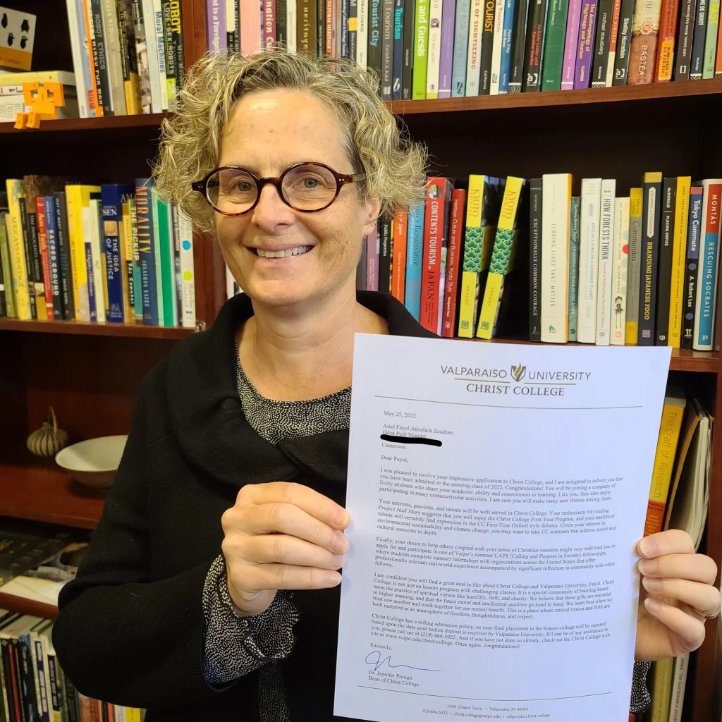With determination, Charles Smith, a smiling person with glasses, holds a letter from Valparaiso University Christ College in front of a bookshelf.