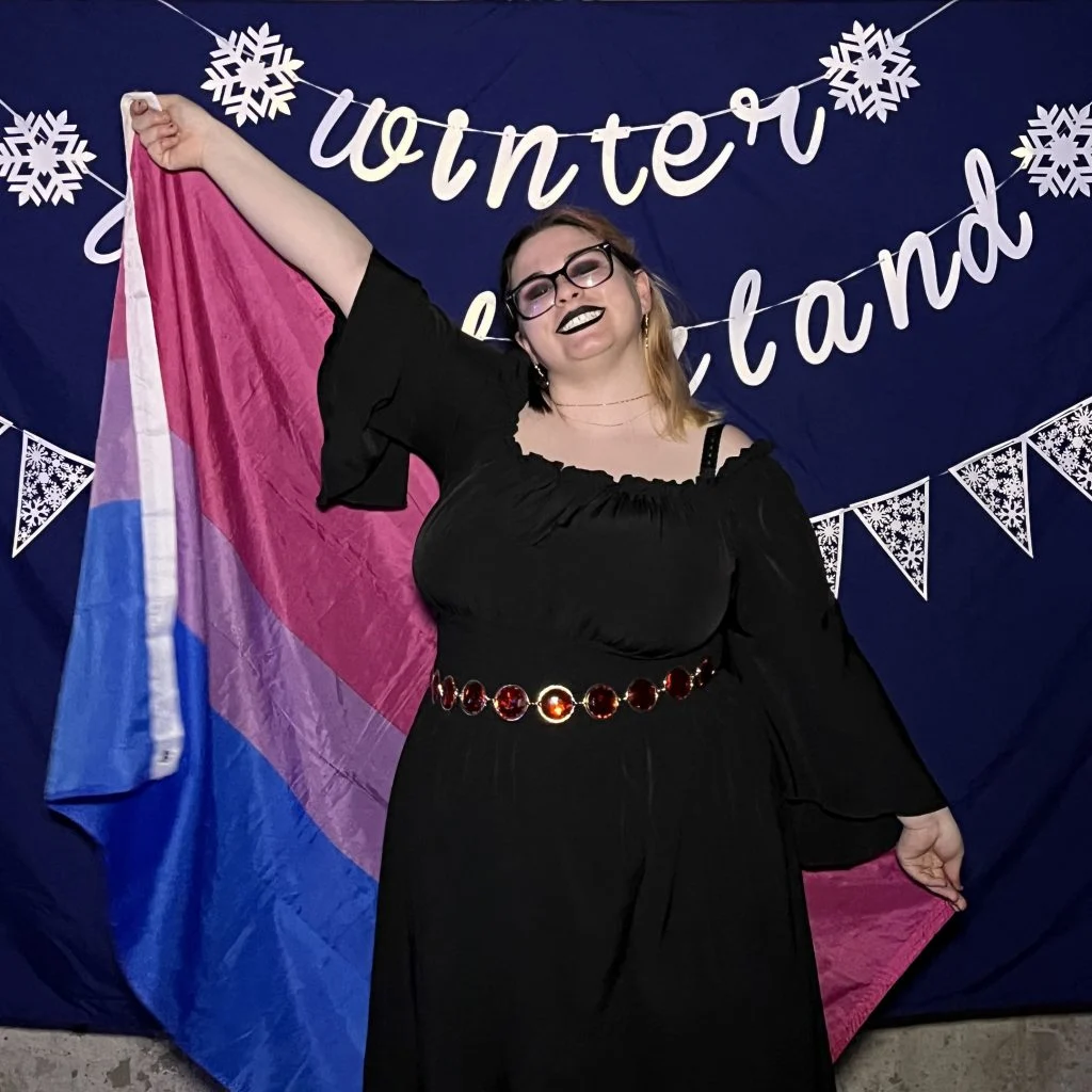 Smiling person in a black dress holding a bisexual pride flag, standing in front of a "Winter Wonderland" banner, bringing light and joy to everyone around them.