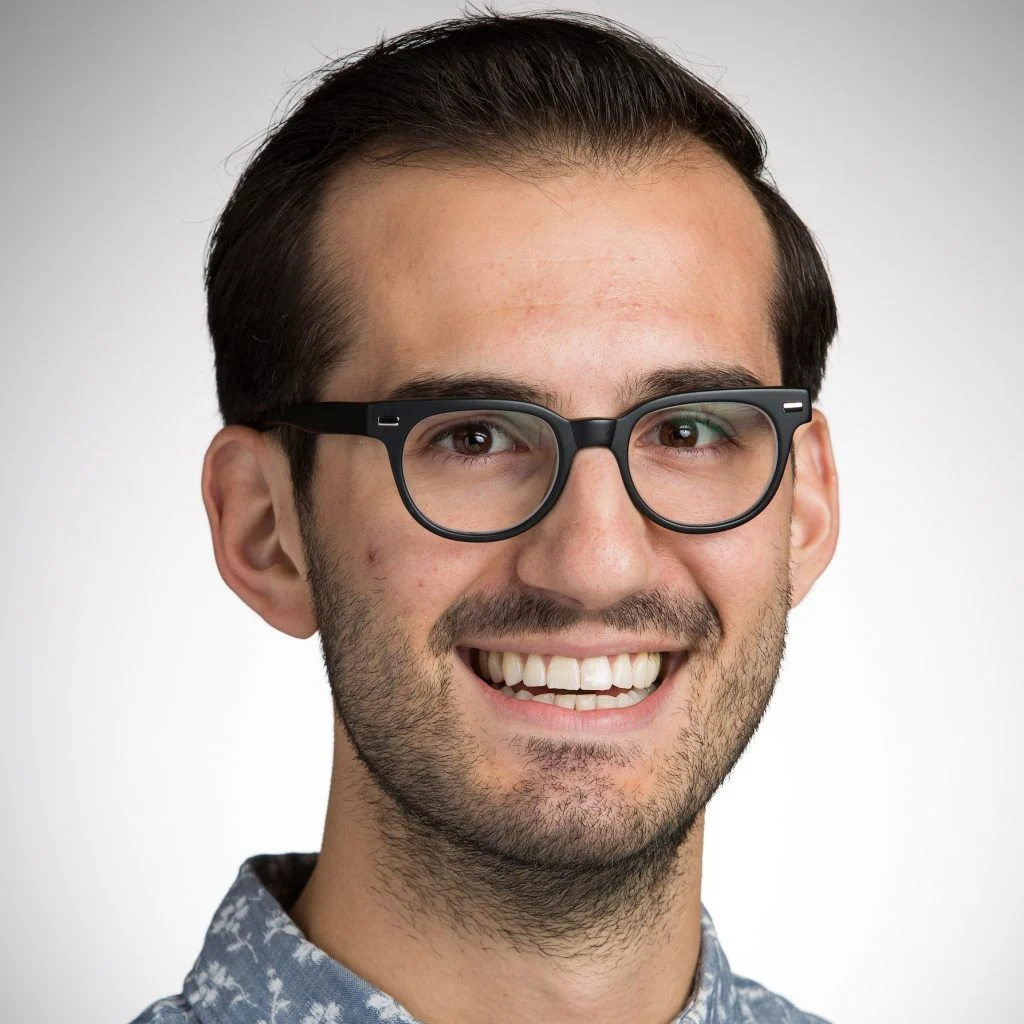 Smiling man with dark hair, glasses, and a patterned shirt stands against a plain light background, radiating warmth that can brighten everyone’s day.