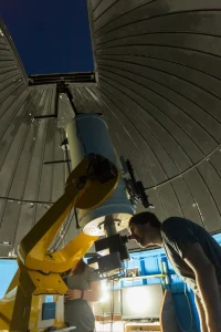 A person peers into a telescope inside a domed observatory with an open roof at night.