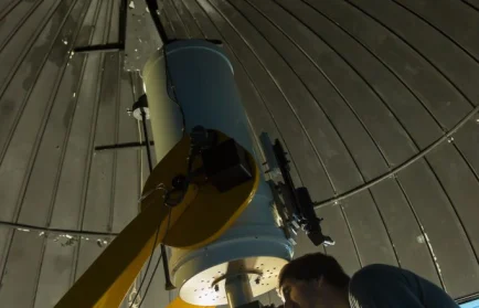 A person peers into a telescope inside a domed observatory with an open roof at night.