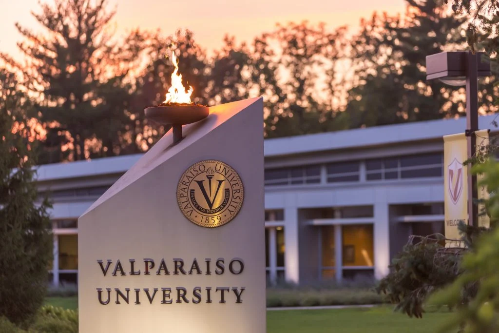 Valparaiso University sign with a burning flame on top, building and trees in the background at sunset, highlighting the campus's Leadership for Public Purpose as recognized by the Carnegie Elective Classification.