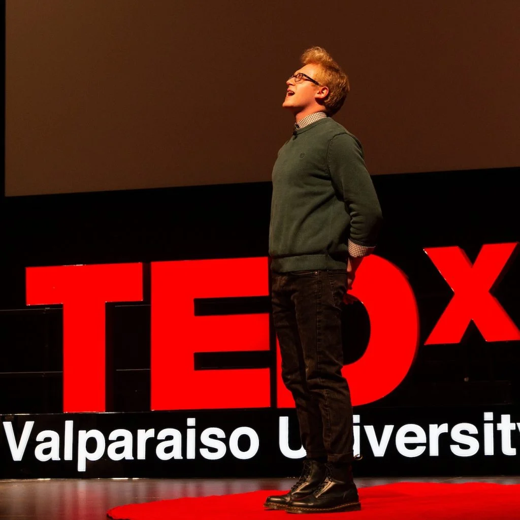 Daniel Owens speaking on stage at TEDx Valparaiso University, standing in front of a large red TEDx sign, sharing insights on Outcomes in Action.