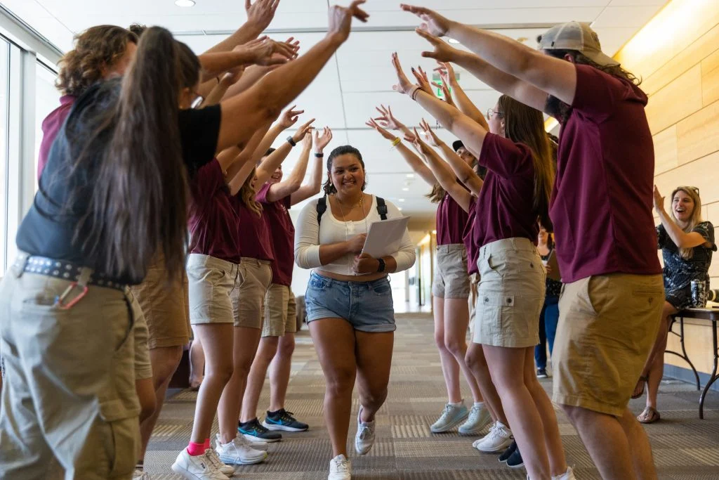 A smiling Jazzy Soria-Gonzalez walks through a tunnel of cheering people with raised arms indoors, celebrating her eventful first year and blooming as a beacon among her peers.