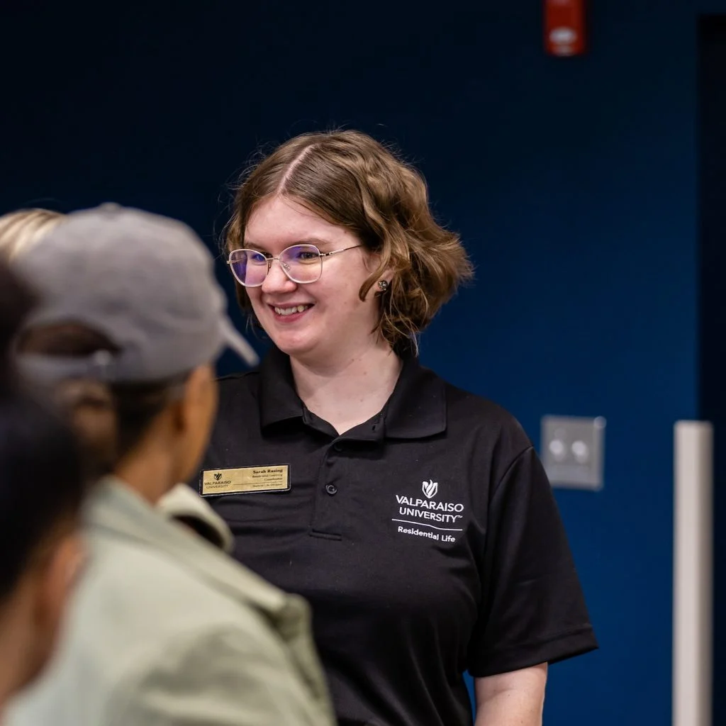 Smiling woman in a Valparaiso University shirt brings light to the room as she talks with everyone indoors against a dark blue wall.