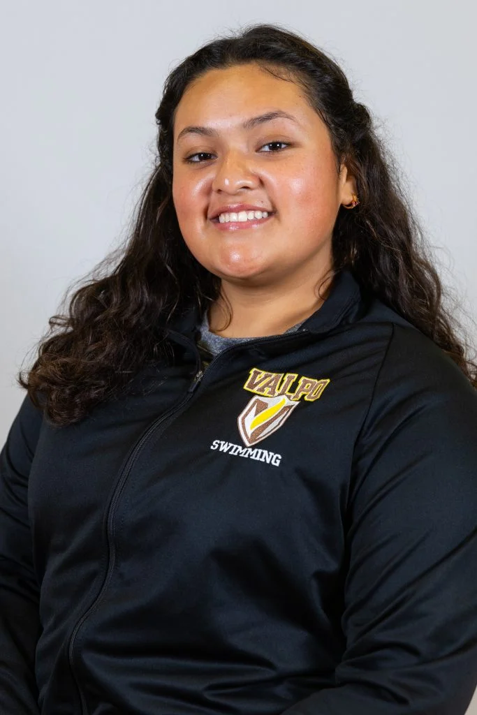 Smiling person with long curly hair, Jazzy Soria-Gonzalez, in a black "Valpo Swimming" jacket, posing against a plain background, capturing the spirit of an eventful first year.