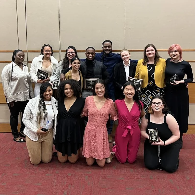 Everyone stands together on a red carpeted floor, some holding plaques, as Light pours in to illuminate the cheerful indoor group.