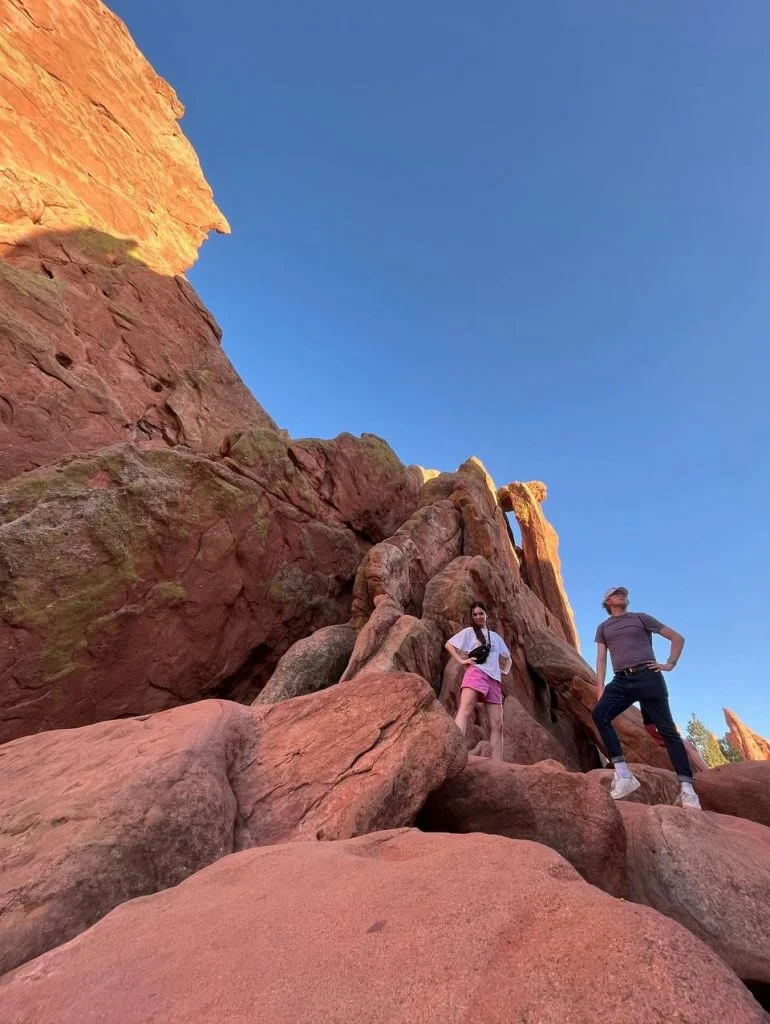 Two people, embodying Outcomes in Action, stand on red rocks under a clear blue sky, surrounded by large rock formations.