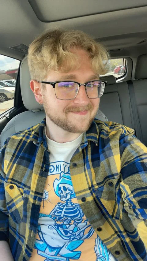 Daniel Owens, with blond hair and glasses, smiles in a car, wearing a yellow flannel and a skeleton graphic t-shirt—capturing an authentic moment from Outcomes in Action 2023.