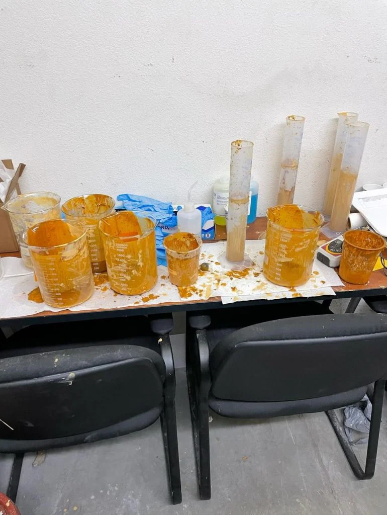 Messy lab table with orange liquid-filled beakers, cylinders, and stains, two black chairs in front—an example of Outcomes in Action inspired by alumni like Daniel Owens.