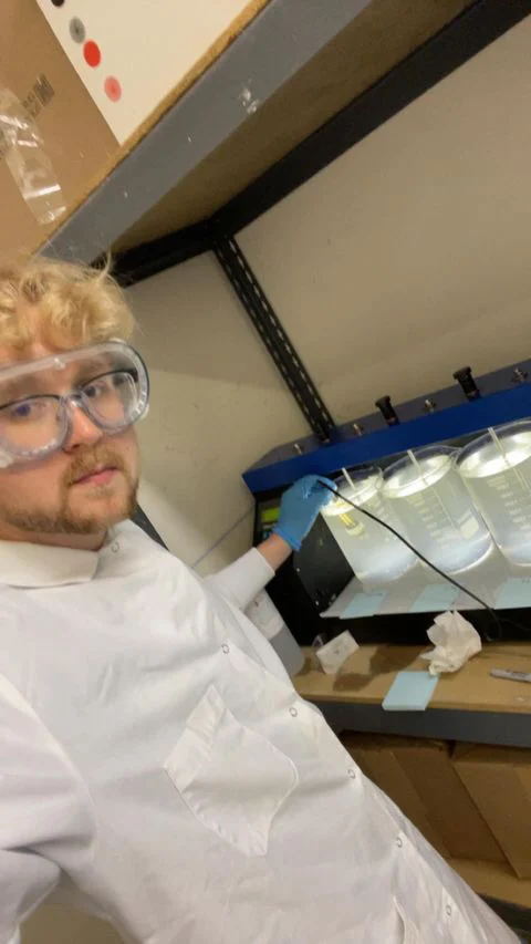 A person in a lab coat and goggles works with equipment under a fume hood in a laboratory, demonstrating Outcomes in Action.