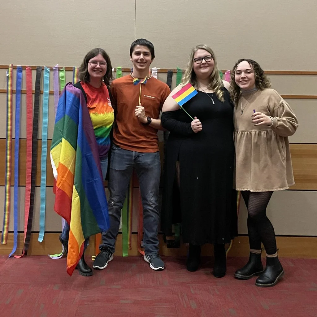 Four people stand smiling indoors, bathed in light, with pride flags and rainbow ribbons behind them, celebrating LGBTQ+ pride with everyone.
