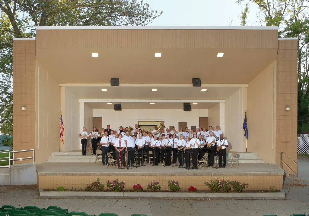 A concert band of Valparaiso University faculty and alumni stands on an outdoor stage, facing the audience with instruments ready to perform inspiring music.