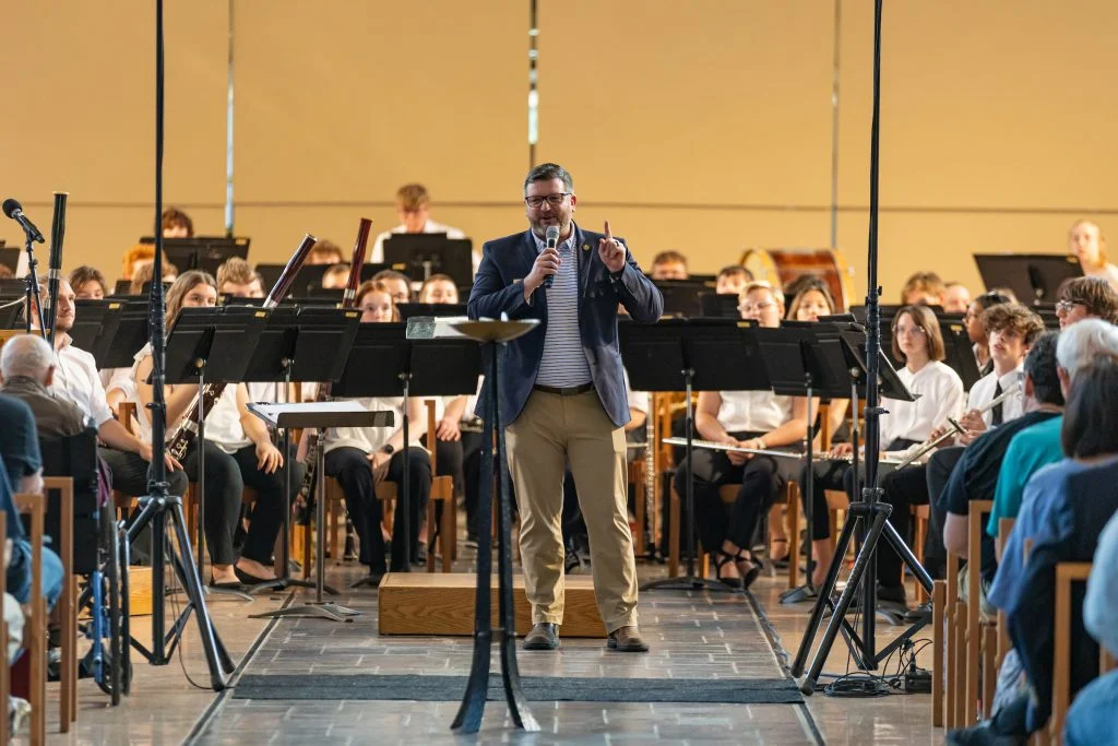 A man speaks to an audience in front of an orchestra seated with music stands in a well-lit hall, celebrating the legacy of Lutheran Summer Music and its liturgical traditions.