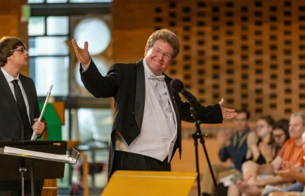 Smiling conductor in a tuxedo gestures to an applauding audience, celebrating the Liturgical Legacy of Lutheran Summer Music in a grand concert hall.