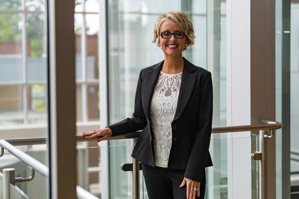 Nadia Paul, smiling in a black suit and glasses, stands by a railing in a modern, bright building—radiating confidence and embodying balanced life and bright outcomes.
