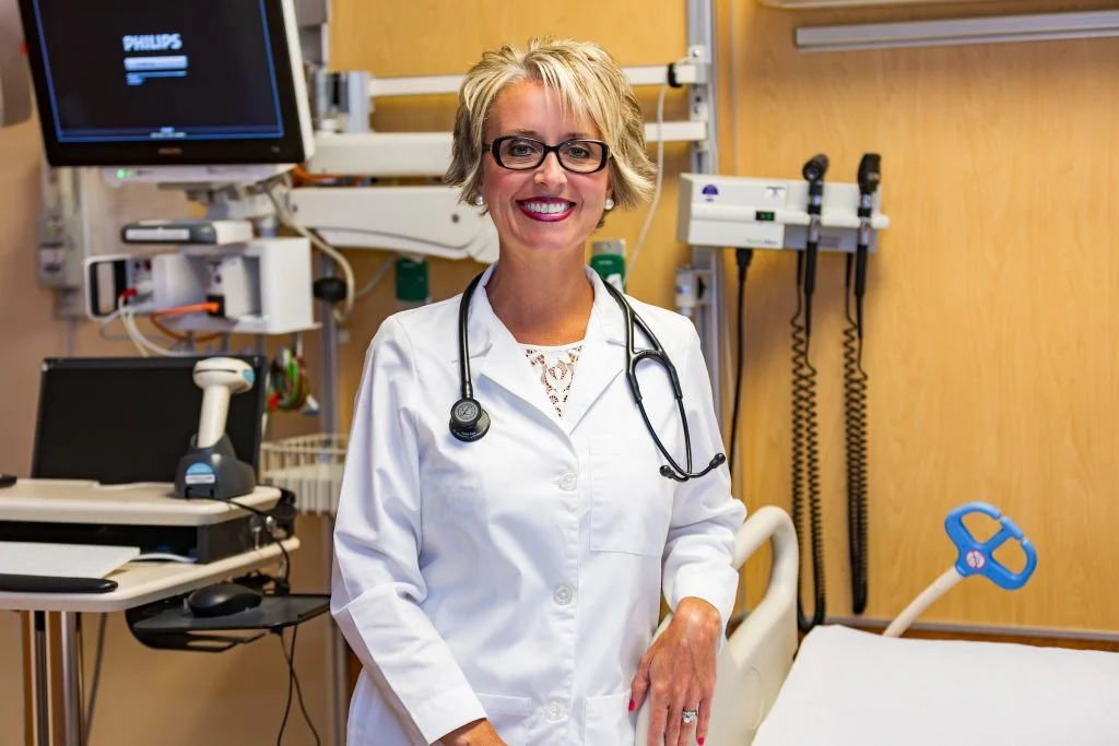 Smiling female doctor Nadia Paul in a white coat stands in a medical exam room with equipment in the background, dedicated to helping patients achieve a Balanced Life and Bright Outcomes.