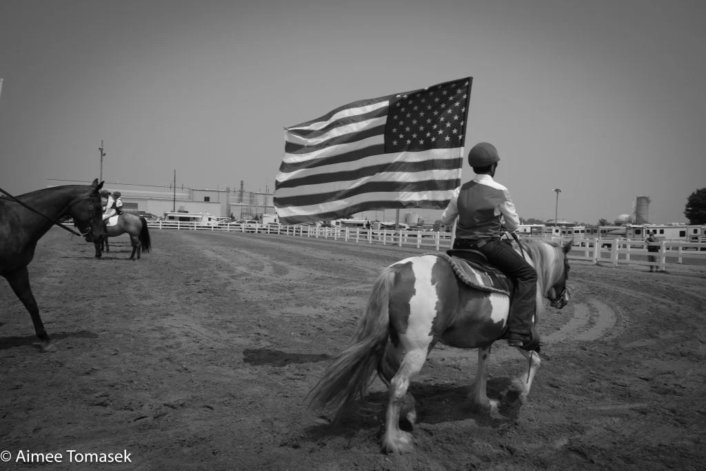 A rider on a pony holds a large American flag in an outdoor arena, with another horse nearby—capturing a scene that inspires future photographers and celebrates the spirit of the local community.