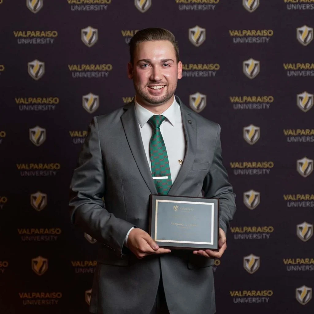 Anthony Luciano, in a suit, holds a plaque while standing before a Valparaiso University backdrop—celebrating Outcomes in Action and inspiring Future Beacons.