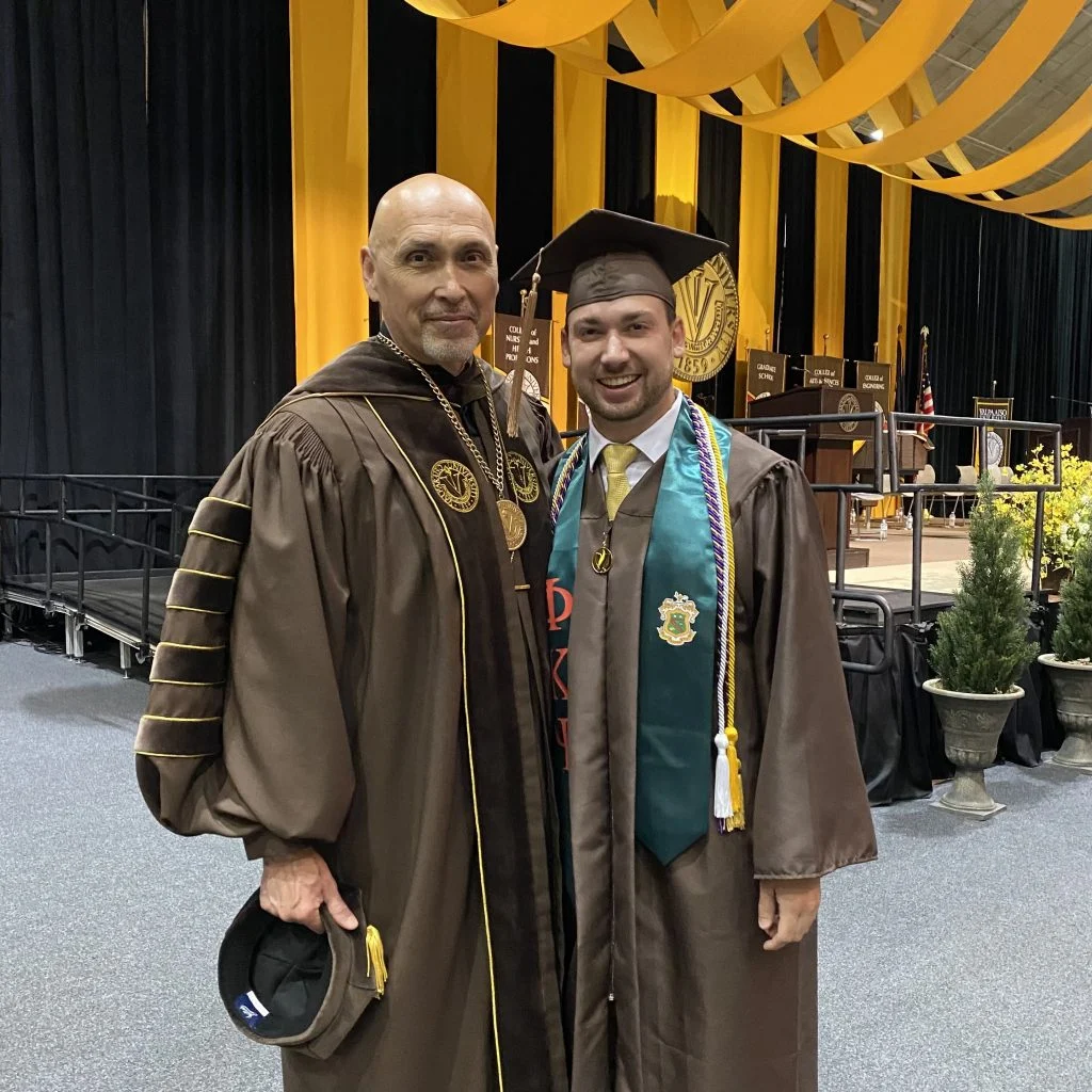 Anthony Luciano and a friend, both in graduation gowns, smile together at a university commencement ceremony with banners behind them—true Outcomes in Action and shining Future Beacons.