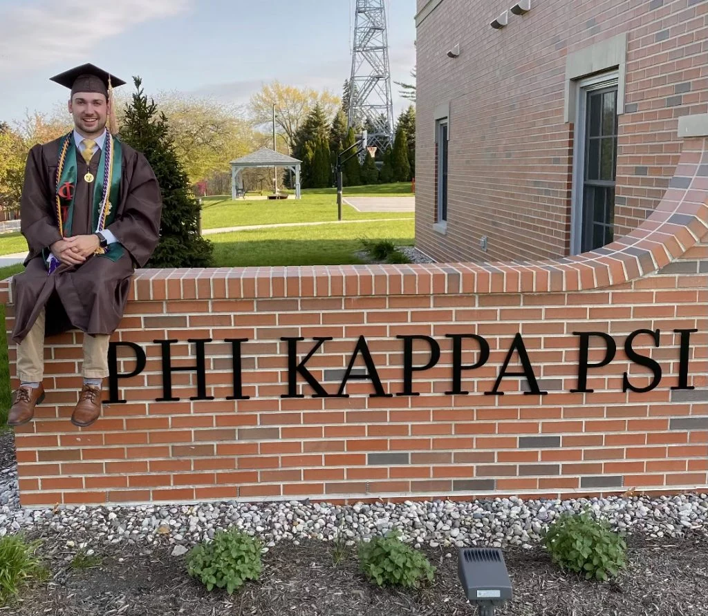 A graduate in cap and gown, embodying Outcomes in Action, sits on a brick wall with "PHI KAPPA PSI" written on it.