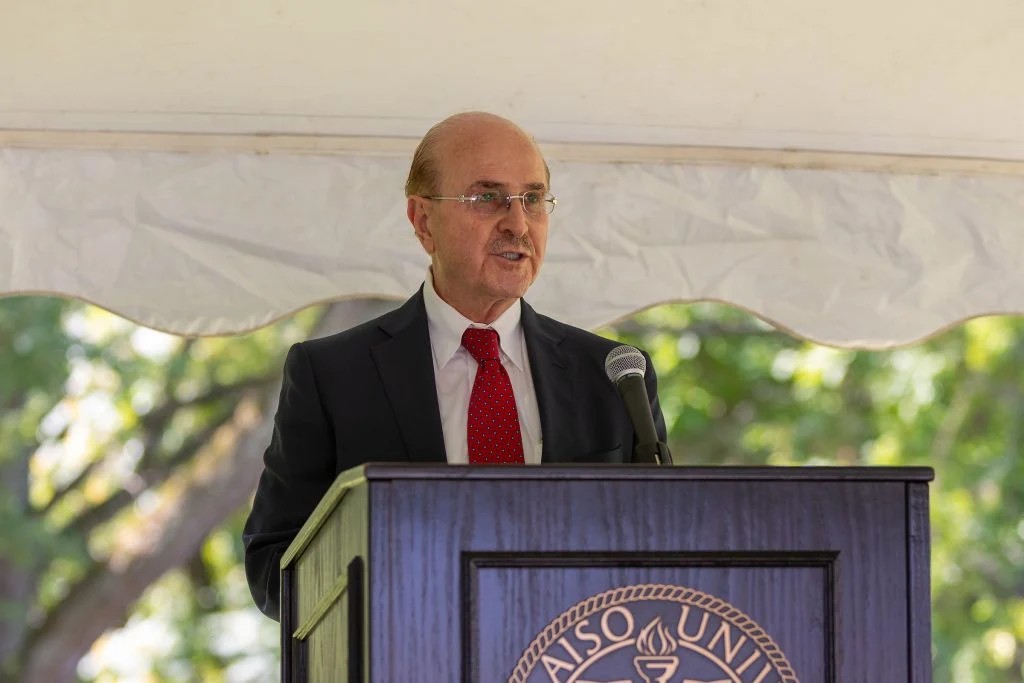 A man in a suit and red tie speaks at a podium outdoors under a white canopy, representing Valparaiso University at the Rifai Institute event.