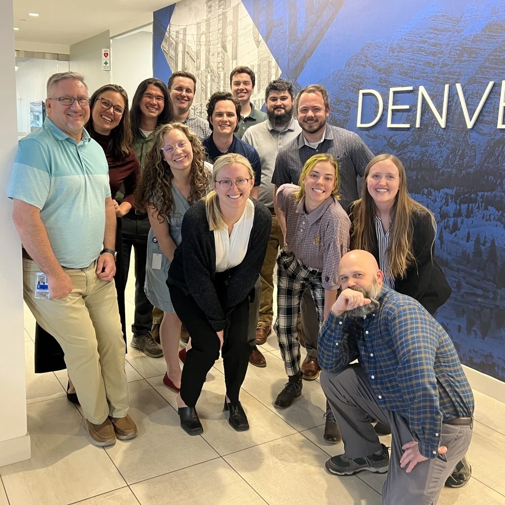 A group of thirteen people, including Electrical Engineer Kallie Lyon, smiling and posing together indoors in front of a wall that says "Denver.