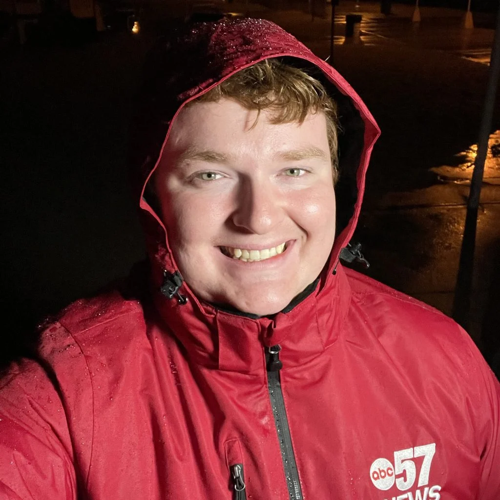 Emmett Twomey, in a red ABC 57 News rain jacket, smiles outdoors at night during a storm, showing dedication to keeping the community informed in wet weather.