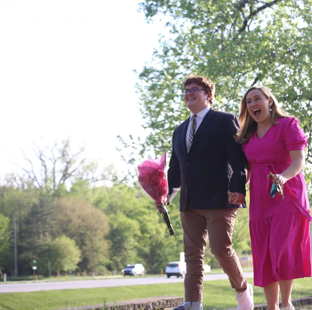 Two people smiling and holding hands outside; one holds flowers and the other, in a bright pink dress, shows how together they can weather any storm.