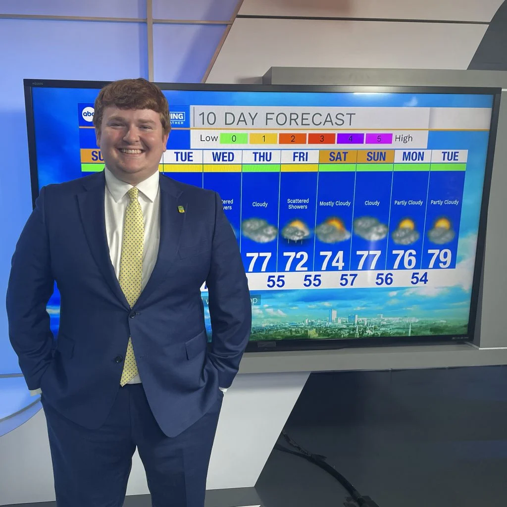 Smiling man in a suit, Emmett Twomey, stands by a screen showing a 10-day weather forecast in a TV studio, ready to share his story with the community.