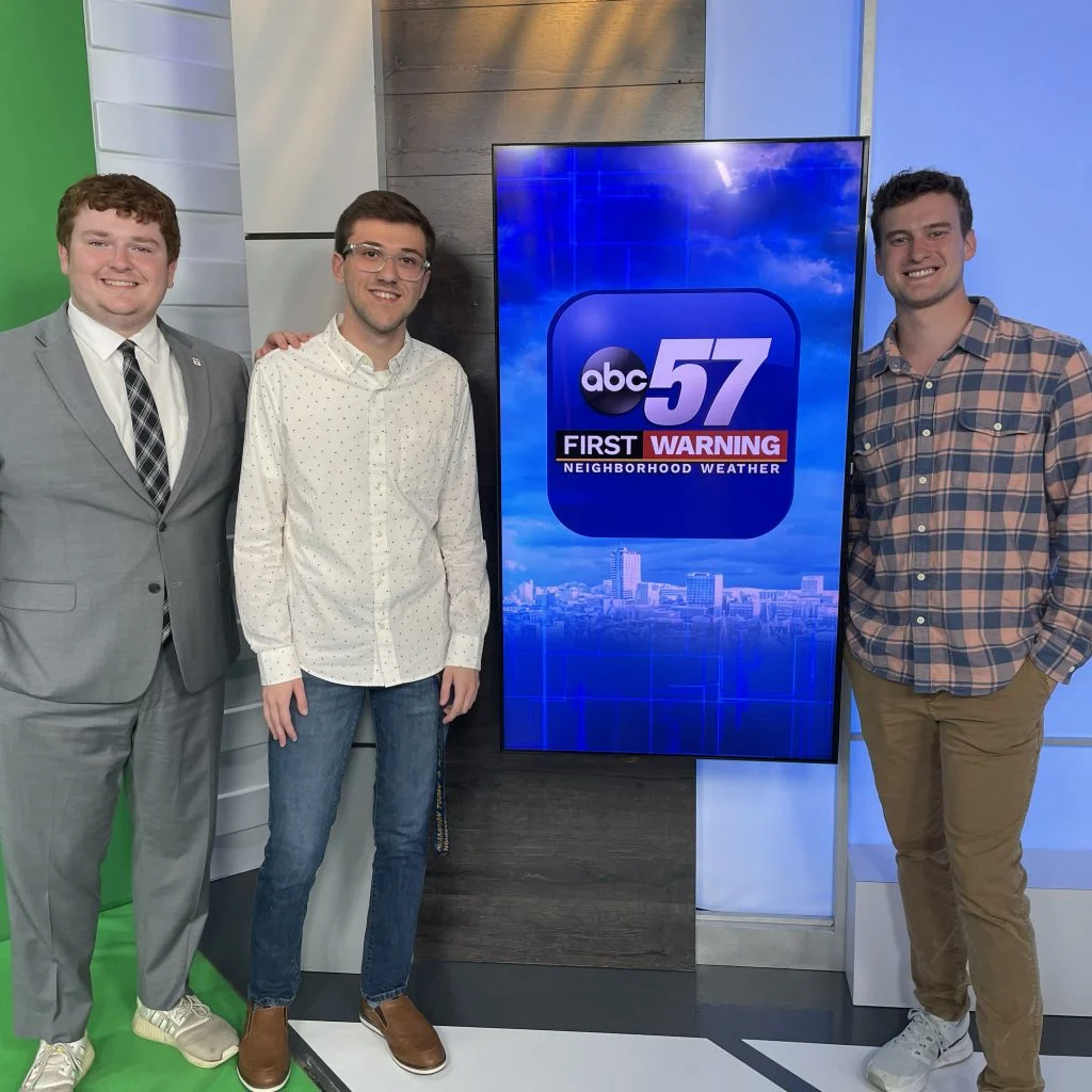 Three men stand smiling beside an “ABC57 First Warning Neighborhood Weather” screen in a studio, highlighting Emmett Twomey’s commitment to keeping the Right Community prepared to Weather Any Storm.