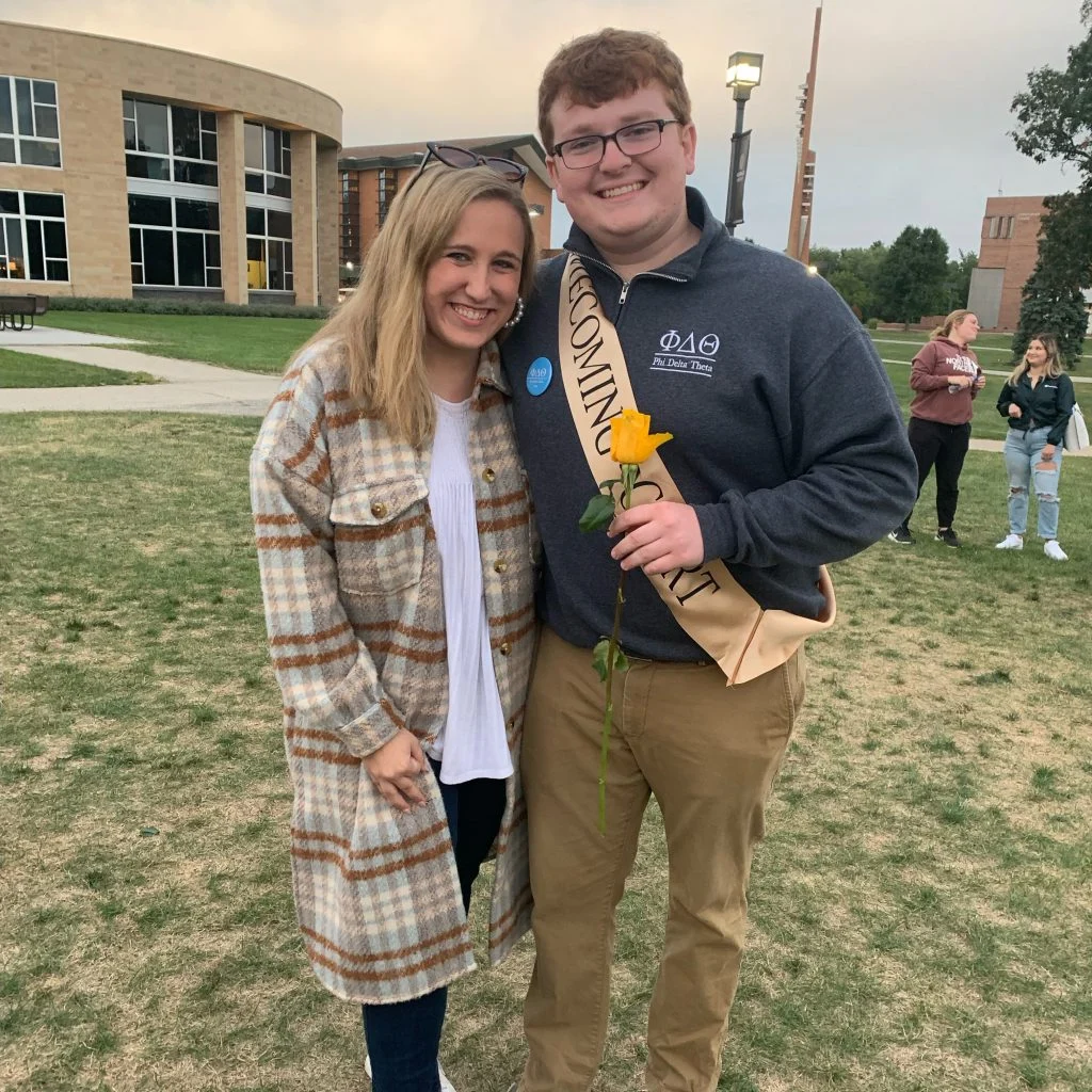 Two smiling people pose outside; one holds a yellow rose and wears a "Homecoming Court" sash, celebrating with Emmett Twomey and showing the strength to Weather Any Storm together.